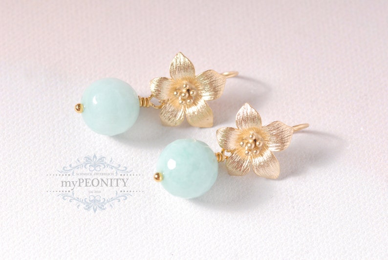 May include: A pair of gold earrings with a light blue round gemstone and a gold flower charm.