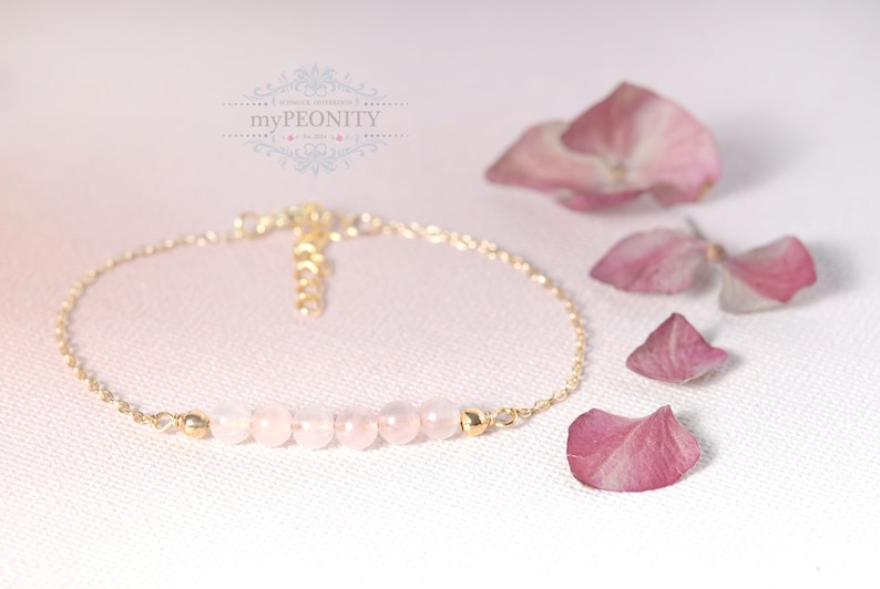 Puede incluir: Un delicado brazalete de cadena dorada con una fila de cuentas de cuarzo rosa p&aacute;lido. El brazalete est&aacute; sobre un fondo blanco con p&eacute;talos de flores rosas.