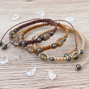 Pode incluir: Quatro pulseiras de macramé em tons de castanho e bege, cada uma com contas transparentes e cinza escuro. As pulseiras estão dispostas sobre uma superfície de madeira clara, com pequenos cristais transparentes espalhados ao redor. As pulseiras são feitas à mão.