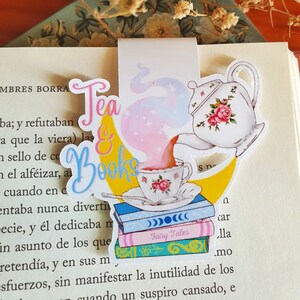 Könnte beinhalten: Eine weiße Teetasse und Teekanne mit rosa Blumen gießen Tee in die Tasse. Die Teetasse steht auf einem Stapel Bücher mit dem Schriftzug "Fairy Tales" auf dem Einband. Der Hintergrund ist ein gelber Halbmond mit einem rosa und violetten Schimmer. Der Text "Tea & Books" ist in einer verspielten Schrift geschrieben.