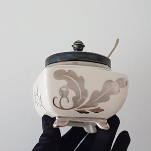 May include: White ceramic sugar bowl with a silver lid and a silver floral design. The bowl has four feet and a small silver spoon.