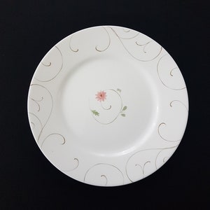 Puede incluir: Un plato de cena de cerámica blanca con un diseño floral dorado y verde. El plato tiene un patrón delicado y en espiral y una sola flor rosa en el centro.