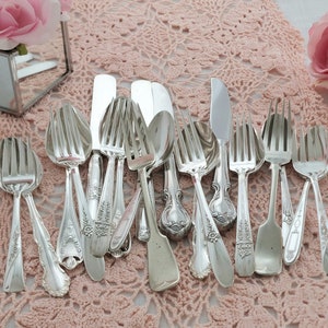 May include: A collection of antique silver cutlery, including forks, spoons, and knives, displayed on a pink lace runner. The silverware features intricate floral and scroll patterns. The text "A Hummingbird Heirloom" is visible.