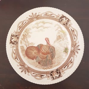 May include: A white ceramic dinner plate with a brown and cream design featuring a turkey in a woodland setting. The plate has a scalloped edge and a decorative border.