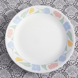 May include: White round plate with a floral border. The border features pastel-coloured tulips in pink, blue, and yellow, with green leaves. The plate is set against a white lace tablecloth.