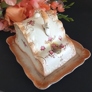 May include: A vintage ceramic butter dish with a lid, featuring a floral design with pink roses and a basket. The dish has a scalloped edge and a handle on top. The color scheme includes cream, peach, and gold accents.