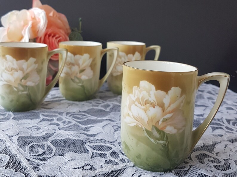 May include: Set of four vintage porcelain teacups with a floral design. The cups are decorated with a white rose on a green and gold background.