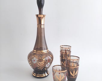 May include: A vintage brown glass decanter set with a tall, slender decanter and six matching glasses. The decanter and glasses feature gold trim and floral designs. The decanter has a dark brown stopper.