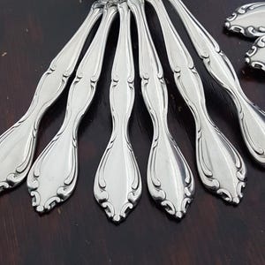 Può includere: Primo piano di diversi cucchiai color argento con manici curvi e ornati. I cucchiai sono disposti a ventaglio, mostrando le loro superfici lucide e bordi decorativi. I cucchiai sono su una superficie marrone scuro.