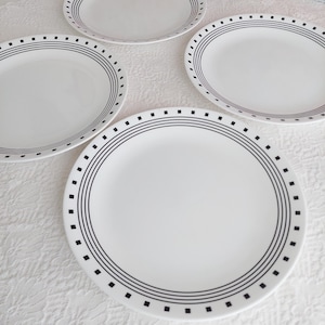 Puede incluir: Cuatro platos de cerámica blanca con un diseño geométrico negro. Cada plato tiene un borde negro con tres líneas paralelas y pequeños acentos cuadrados. Los platos están dispuestos sobre una superficie blanca texturizada.