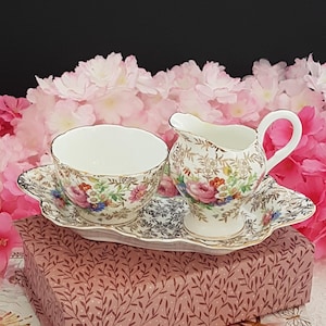 以下が含まれることがあります： ティーカップ、クリーマー、サービングトレイを備えたヴィンテージティーセット。セットは白地に花柄と金色のアクセント。トレイの長さは約25cmです。