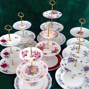 May include: A selection of vintage cake stands with gold-tone handles. The plates display floral designs in shades of pink, red, and purple, set against a white backdrop. The stands are displayed on a green surface.