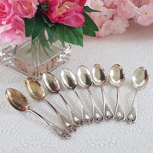 May include: A collection of antique silver spoons arranged on a white lace tablecloth. The spoons have ornate handle designs and are displayed near a vase of pink flowers. The spoons are a vintage silverware set.