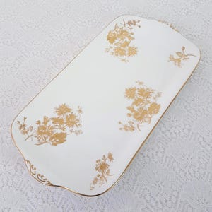 May include: A white rectangular serving platter with gold floral patterns and a gold trim. The tray has rounded corners and a slightly raised edge. The floral design is repeated across the surface.