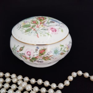 May include: A white porcelain trinket box with a floral design. The lid and base of the box are decorated with a colourful floral pattern featuring pink, blue, and green flowers. The box has a gold trim around the top edge.