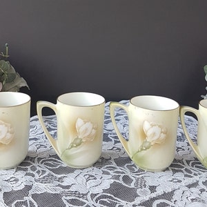 May include: Set of four vintage porcelain teacups with floral designs. Each cup features a delicate white flower with green leaves on a pale yellow background.