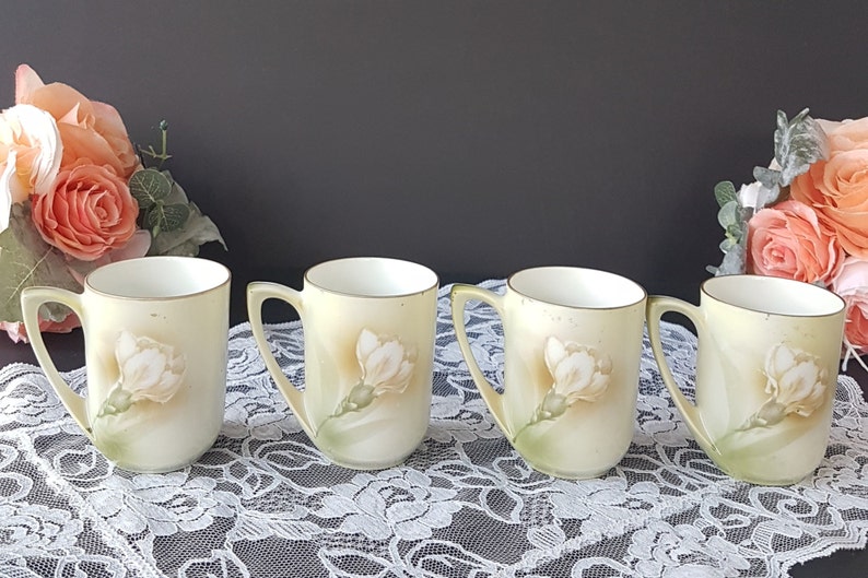 May include: Set of four vintage porcelain teacups with floral designs. Each cup features a delicate white flower with green leaves on a pale yellow background.