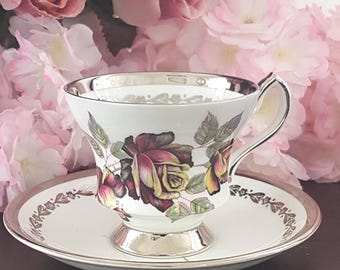 May include: A white porcelain teacup and saucer set featuring a detailed illustration of dark red and yellow roses. The rim of the cup, the base, and the edge of the saucer are finished with a metallic silver trim and a floral pattern.