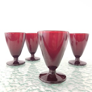 May include: Four red glass goblets with a wide base and a flared top. The goblets are arranged in a row on a textured surface.
