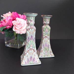 May include: Two decorative ceramic candle holders with a floral design in shades of purple, green, and pink. The candle holders have a square base and a flared shape. The tops are a light green color.