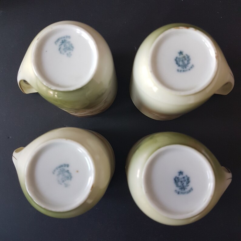 May include: Four white porcelain teacups with green and gold floral designs. The bottom of each cup has a blue and white logo with the text "Germany" in cursive.