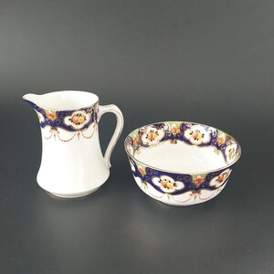 May include: A white porcelain creamer and sugar bowl set featuring a deep blue border with gold filigree, orange and yellow floral motifs, and delicate gold-toned swags. Both pieces have a smooth finish and a curved, elegant silhouette.