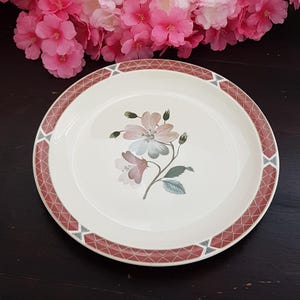 May include: A round, off-white ceramic plate with a floral design. The plate has a border of red geometric shapes and a central illustration of pink and blue flowers with green leaves. A decorative piece for serving.