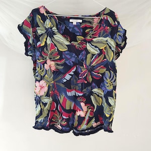 May include: A navy blue, short-sleeved top with a V-neck and a vibrant floral pattern. The design features large leaves and flowers in shades of pink, red, green, and yellow. The top has a fringed trim along the sleeves and hem.