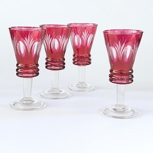 May include: Four matching stemmed glasses featuring a deep cranberry red upper bowl with clear cut glass leaf and oval patterns. Each glass has a clear glass stem with three horizontal rings above the base and a clear circular foot.