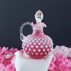 Könnte beinhalten: Ein rosa Hobnail-Glaskrug mit einem durchsichtigen Griff und Stopfen. Der Krug ist mit weißen Hobnails verziert und hat einen ausgestellten Rand. Der Stopfen ist klar und hat ein strukturiertes, konisches Design. Der Krug wird von einem weißen Handschuh gehalten.