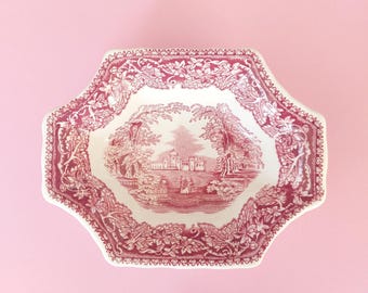 May include: A cream-colored ceramic bowl with a scalloped edge and a deep pink transferware pattern. The center features a landscape with a building and figures, while the border displays a detailed floral and vine motif in matching pink.
