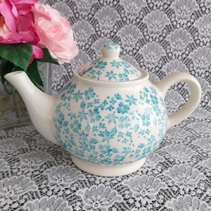 May include: A white ceramic teapot with a lid, adorned with a pattern of small, light blue flowers. The teapot has a curved spout and handle, and sits on a small base. Pink flowers are visible in the background.