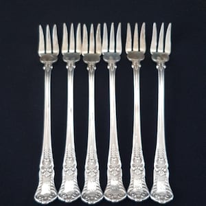 May include: Six silver forks with ornate handles. The forks are arranged in a row, with the tines facing up.