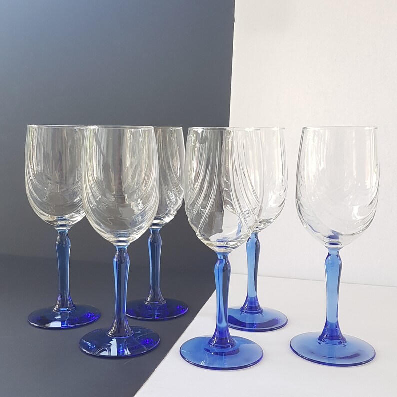 May include: Six clear glass wine glasses with blue stems. The glasses are arranged in a row on a white surface.