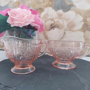 May include: A pink glass creamer and sugar bowl set. The creamer has a spout and handle, while the sugar bowl has two handles. Both pieces feature a floral pattern and are set on a dark surface. A vase of pink flowers is in the background.