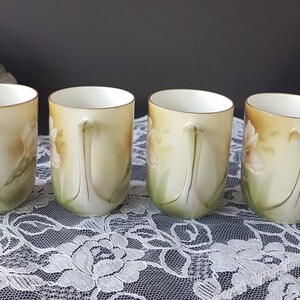 May include: Four vintage porcelain teacups with floral designs. The cups are white with a yellow, green, and pink floral pattern. The cups have gold trim around the rim and handles.
