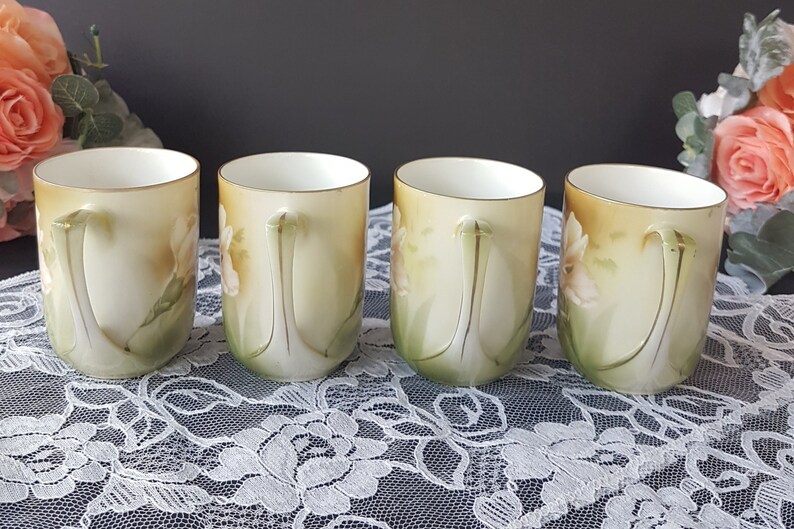 May include: Four vintage porcelain teacups with floral designs. The cups are white with a yellow, green, and pink floral pattern. The cups have gold trim around the rim and handles.