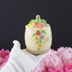 Könnte beinhalten: Ein cremefarbenes Keramikgefäß mit Deckel, verziert mit bunten Blumen und grünen Blättern. Das Gefäß hat eine Korbgeflechtstruktur und wird von einer behandschuhten Hand gehalten. Der Deckel hat einen blütenförmigen Griff.