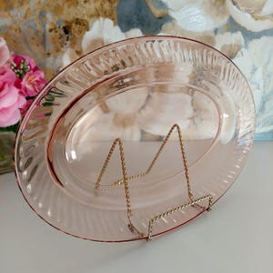 May include: A vintage, oval pink glass serving platter with a textured rim, displayed on a gold-coloured metal stand. The background features a floral arrangement and a patterned wall.