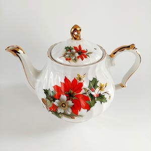 May include: White porcelain teapot with a gold handle, spout, and lid accents. The teapot is decorated with red poinsettias, white flowers, and green holly leaves. The lid has a gold finial.