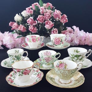 May include: A collection of vintage teacups and saucers with floral designs, including roses and other flowers. The teacups are white, green, and gold, with delicate patterns. A bouquet of pink roses is in the background.