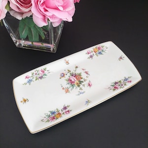 Peut inclure: Un plateau de service rectangulaire en céramique blanche avec un motif floral. Le plateau présente une bordure dorée et est orné de grappes de fleurs colorées roses, jaunes, bleues et violettes. Les fleurs sont disposées symétriquement.