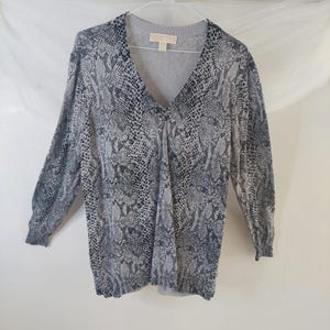 May include: A long-sleeved sweater with a V-neck and a snakeskin pattern in shades of gray and black. The sweater is hanging on a white hanger. The brand name "Michael" is visible on the label.