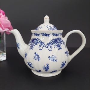 May include: White ceramic teapot with a blue floral design. The teapot has a curved spout, a rounded body, and a matching lid with a small gold accent. The handle is a large, curved loop. The floral pattern includes small and large flowers.