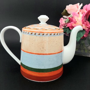 Op de afbeelding: Een keramische theepot met een witte handgreep en tuit. De body heeft horizontale strepen in oranje, blauw en groen, met een beige gespikkelde achtergrond. Het deksel is wit met een decoratieve knop en een patroonrand.