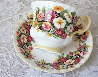 May include: White teacup and saucer set decorated with a floral pattern in shades of pink, yellow, and white. The teacup has a yellow handle and base, and the saucer has a matching floral border. The set is placed on a white lace tablecloth.