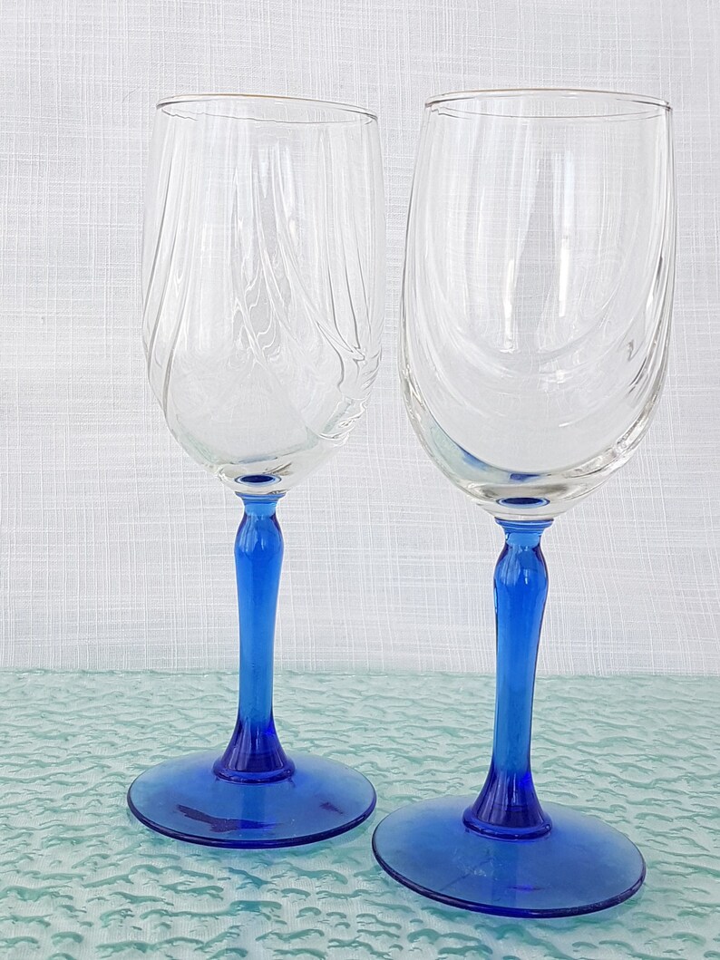 May include: Two clear glass wine glasses with blue stems and bases. The glasses are empty and sitting on a blue surface.