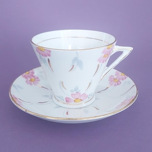 May include: A white porcelain teacup and saucer set featuring a delicate pattern of pink flowers with yellow centres, accented by light blue leaves and thin gold trim along the rims and handle. The cup has a flared shape and a triangular handle.