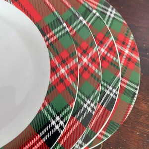 Könnte beinhalten: Ein Stapel weißer Keramikteller mit einem rot, grün, schwarz und weiß karierten Muster am Rand. Die Teller sind leicht überlappend angeordnet und zeigen das sich wiederholende Design. Die Teller dienen wahrscheinlich zum Servieren von Speisen.