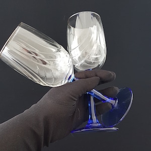 May include: Two clear glass wine glasses with blue stems and bases. The glasses have a swirl design and are held by a black gloved hand.